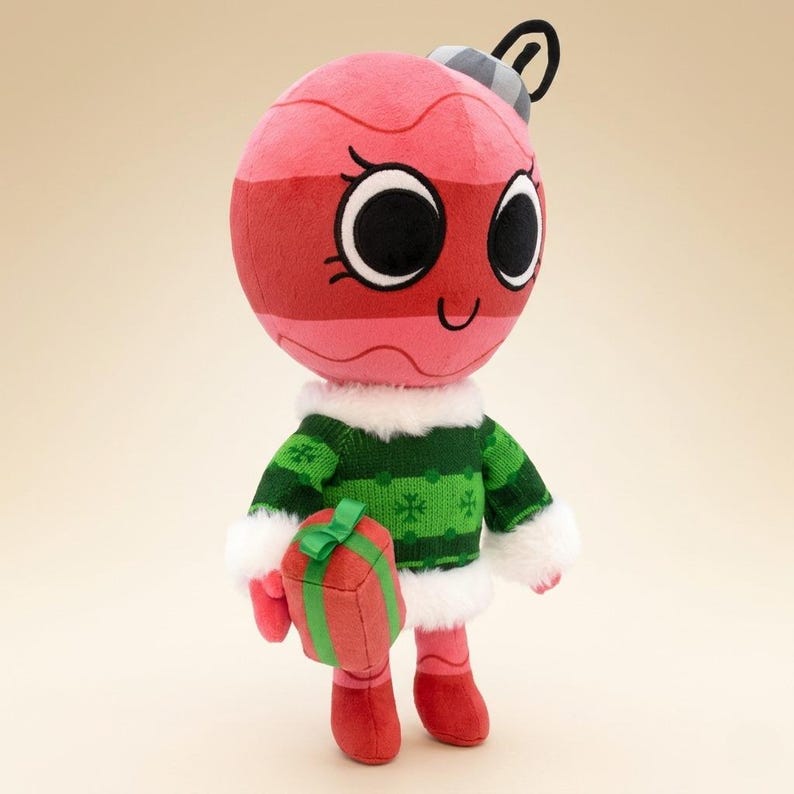 K&ouml;nnte beinhalten: Ein Pl&uuml;sch-Weihnachtsschmuck-Charakter mit einem l&auml;chelnden Gesicht, der einen gr&uuml;nen Pullover mit wei&szlig;em Besatz tr&auml;gt und ein rotes Geschenk h&auml;lt. Der Schmuck ist rot und rosa mit schwarzen Augen und einem silbernen Oberteil.
