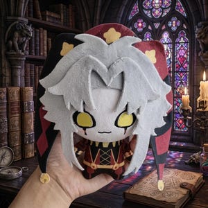 Könnte beinhalten: Plüschpuppe einer Figur mit weißen Haaren, gelben Augen und einer Narrenkappe. Die Puppe trägt ein rot-schwarzes Outfit. Der Hintergrund umfasst ein Buntglasfenster, Kerzen und ein Bücherregal.