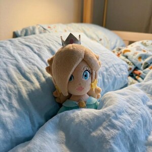 May include: A plush doll of a character with blonde hair, a silver crown, and a teal dress, resting on a bed with light blue bedding. The doll has blue eyes and star-shaped earrings.