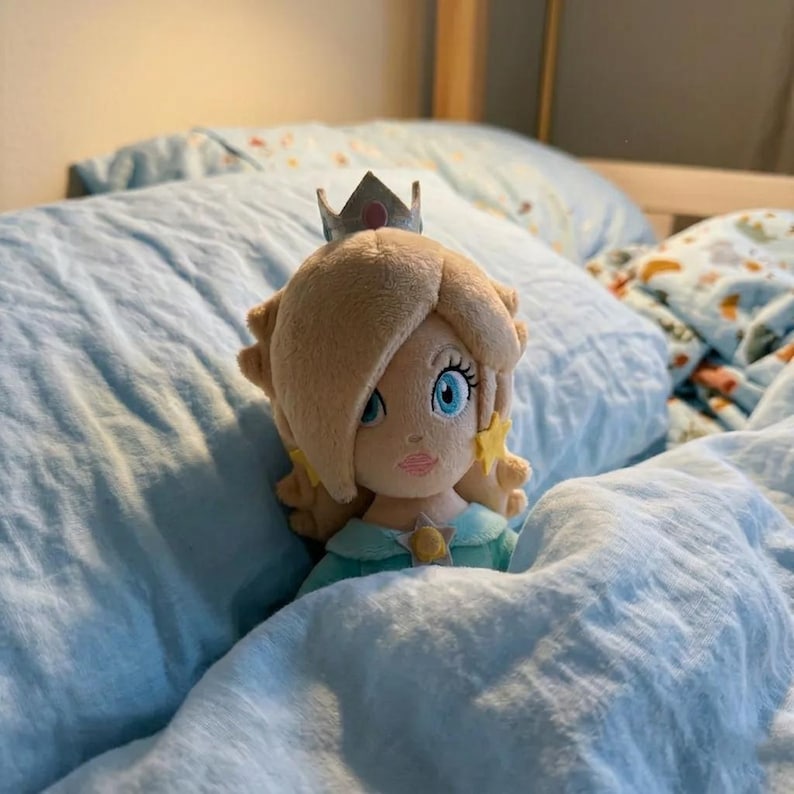 May include: A plush doll of a character with blonde hair, a silver crown, and a teal dress, resting on a bed with light blue bedding. The doll has blue eyes and star-shaped earrings.