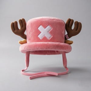 以下が含まれることがあります： 前面に大きな白い刺繍のXが施されたピンクのぬいぐるみハット。両サイドには茶色の鹿の角が突き出ており、つばからは2本の長いピンクの布製ストラップが垂れ下がっています。帽子全体が柔らかくふわふわとした質感です。