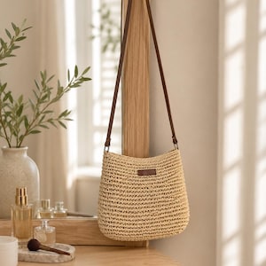May include: A beige woven straw crossbody bag with a long, thin brown leather strap. The bag features a rectangular brown leather patch centred on the front upper portion. The texture of the straw is visible in a horizontal, repeating pattern.