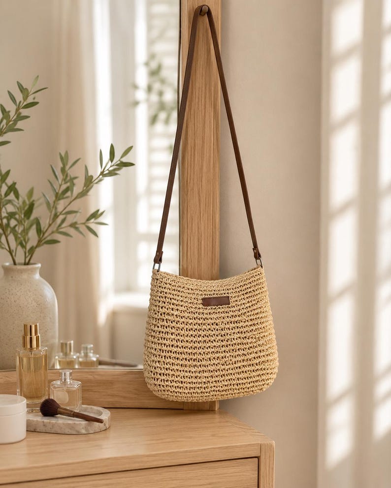 May include: A beige woven straw crossbody bag with a long, thin brown leather strap. The bag features a rectangular brown leather patch centred on the front upper portion. The texture of the straw is visible in a horizontal, repeating pattern.
