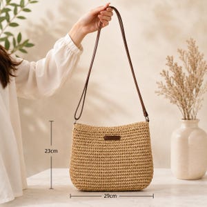May include: A tan woven straw shoulder bag with a dark brown leather-like strap and a small rectangular brown patch on the front. The bag measures 23 cm in height and 29 cm in width, featuring a textured, open-weave construction throughout.