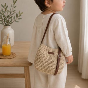 May include: A beige woven straw crossbody bag with a brown leather-look strap and a small rectangular brown patch on the front. The bag features a silver-toned metal buckle on the strap for length adjustment and a simple, structured design.