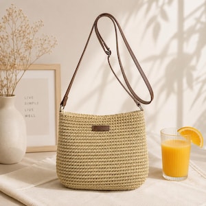 May include: A beige woven straw crossbody bag with a brown adjustable shoulder strap and a small brown rectangular patch on the front. The bag features a textured, knit-like surface and a simple, structured shape with a flat base and rounded corners.