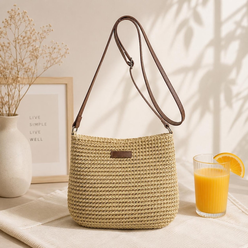 May include: A beige woven straw crossbody bag with a brown adjustable shoulder strap and a small brown rectangular patch on the front. The bag features a textured, knit-like surface and a simple, structured shape with a flat base and rounded corners.