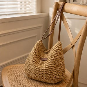 May include: A beige hobo bag crafted from woven material with a slouchy silhouette. The bag features two thin brown leather straps and a small rectangular brown leather patch centred on the front. The texture is consistent and open-knit.