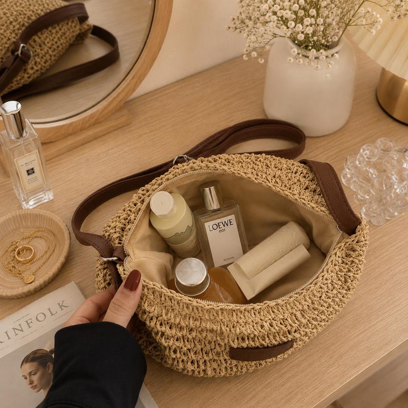 May include: A round woven straw crossbody bag with a dark brown leather strap and trim. The interior is lined with beige fabric and contains a Loewe 001 perfume bottle, a white cylindrical container, and a small amber glass bottle with a silver cap.