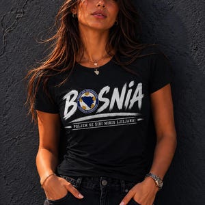 Op de afbeelding: Een zwart t-shirt met korte mouwen met het woord BOSNIA in witte penseelstreekletters, waarbij het logo van de voetbalbond van Bosnië en Herzegovina de O vervangt en de tekst POLJEM SE ŠIRI MIRIS LJILJANA eronder is gedrukt.