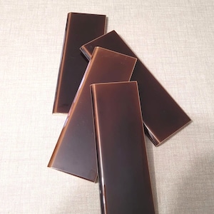 May include: A collection of four rectangular ceramic tiles with a glossy, dark brown finish. Each tile features a smooth, reflective surface and clean, straight edges, arranged in an overlapping, layered pattern to showcase the deep colour.