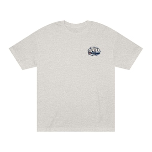 May include: Pale grey t-shirt with a small graphic on the upper left chest. The graphic features the words "Cloud Surfer" in a stylized font, with a wave and surfboard design in blue and white.