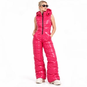 Puede incluir: Un mono sin mangas de color rosa intenso con un acabado acolchado brillante, cuello alto con capucha incorporada y cierre de cremallera central en negro. La prenda presenta un cuerpo ajustado y pantalones anchos, combinados con zapatos blancos.