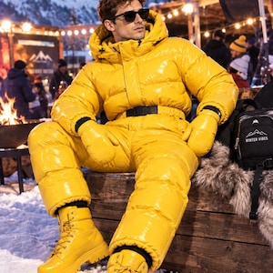 Op de afbeelding: Een felgeel, hoogglanzend, geïsoleerd skipak uit één stuk met een capuchon, een zwarte tailleriem en bijpassende gele wanten. De outfit wordt gecombineerd met gele winterlaarzen met veters en dikke profielzolen. Het pak heeft een glanzende afwerking.