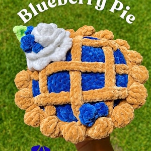 Puede incluir: Un pastel de arándanos tejido a ganchillo con una corteza de rejilla color canela y relleno azul oscuro. Presenta un copete de nata blanca decorado con pequeñas bayas azules y una hoja verde. El texto "Blueberry Pie" aparece en blanco arriba.