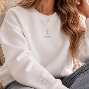 Könnte beinhalten: Ein weißes Sweatshirt mit Rundhalsausschnitt, das ein kleines, mittig platziertes graues Kreuzsymbol über dem Wort BLESSED zeigt. Auf dem linken Ärmel steht der Text I AM BLESSED Psalm 1:1 in grauer Schrift. Das Kleidungsstück hat lange Ärmel.