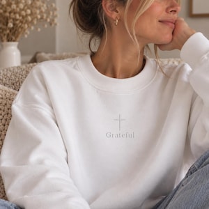Könnte beinhalten: Ein weißes Sweatshirt mit Rundhalsausschnitt und einem kleinen, mittig platzierten Stickmotiv auf der Brust. Das Design besteht aus einem dünnen, vertikalen Kreuzsymbol direkt über dem Wort Grateful, beides in hellgrauem Garn gestickt.