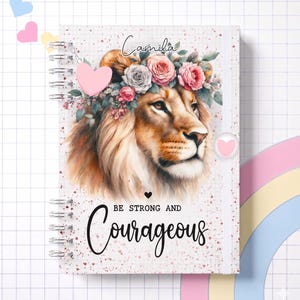 Pode incluir: Um caderno espiral com uma ilustração da cabeça de um leão adornada com uma coroa de flores. A capa inclui o nome "Camila" e as palavras "Be Strong and Courageous". Um pingente de coração rosa está presente.