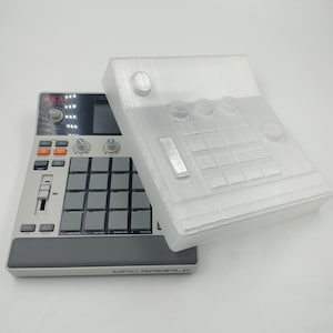 Puede incluir: Un sampler MPC gris y negro con una cubierta de plástico transparente. El sampler tiene una cuadrícula de pads, botones y perillas grises. La cubierta está a un lado, revelando los controles. El texto "MPC SAMPLE" es visible en la parte inferior.