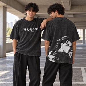 Può includere: Una t-shirt nera lavata all'acido con testo giapponese bianco e il nome inglese EREN YEAGER sul petto. Il retro della maglietta mostra una grande e dettagliata illustrazione grafica bianca di un personaggio dai capelli scuri con un'espressione concentrata.
