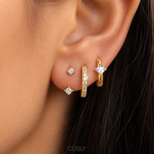Puede incluir: Un conjunto de cuatro pendientes dorados usados en una sola oreja. La colección incluye dos pequeños pendientes de botón con piedras claras, un pequeño aro con piedras claras y un aro con una piedra clara de corte cuadrado en el centro.