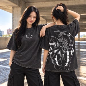 May include: A charcoal grey acid-wash t-shirt featuring a small white graphic on the chest and a large, detailed white illustration on the back depicting a character with hands positioned in a specific gesture, styled with short sleeves and a crew neck.