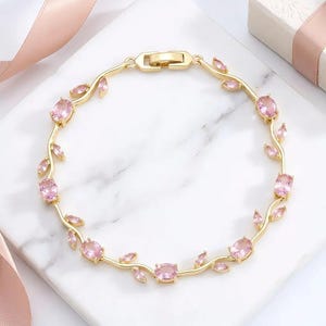 May include: A gold bracelet with pink gemstones in oval and leaf shapes. The bracelet has a delicate, vine-like design and a clasp. The jewellery is displayed on a white marble surface.