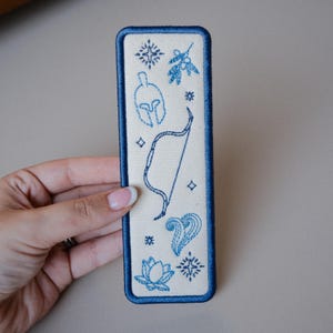 May include: A rectangular fabric bookmark with a cream-coloured base and a dark blue stitched border. The surface features embroidered blue line art designs including a Spartan helmet, a bow, a lotus flower, a sprig, and various star-like symbols.