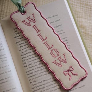 May include: A cream-coloured fabric bookmark with a scalloped edge finished in pink embroidery thread. The bookmark features the name Willow stitched in pink cross-stitch lettering, a small pink bow icon, and a green ribbon loop at the top.
