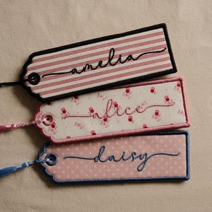 May include: Three rectangular fabric bookmarks with rounded corners and stitched edges. The top bookmark has red stripes and the name amelia. The middle one features a floral pattern and the name alice. The bottom one has pink dots and the name daisy.