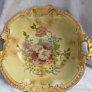 Könnte beinhalten: Antike Servierplatte aus Porzellan mit floralem Design. Die Platte hat einen gewellten Rand und goldfarbene Verzierungen. In der Mitte befindet sich ein Bouquet aus rosa, weißen und blauen Blumen. Zwei goldene Griffe befinden sich auf beiden Seiten der Platte.