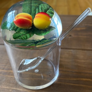 May include: A clear plastic cup with a clear plastic spoon. Inside the cup is a glass dome with two peach-shaped objects and green leaves. The peaches are yellow and red. The spoon is resting on the side of the cup.