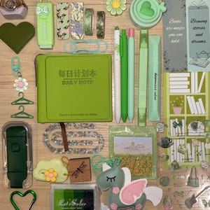 May include: A collection of green stationery items including a Daily Note planner, pens, highlighters, washi tape, a unicorn keychain, a heart-shaped case, a dragonfly stamp, stickers, and bookmarks with the text Books are magic you can hold.
