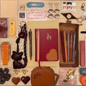 May include: A collection of stationery items including a red notebook with a heart, various pens, stickers with autumn themes, bookmarks, a brown pencil case, a small brown cat-shaped pouch, paper clips, and small decorative charms on a surface.