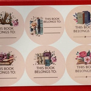 K&ouml;nnte beinhalten: Bogen mit sechs runden Aufklebern mit Blumen- und Buchillustrationen. Jeder Aufkleber tr&auml;gt den Text "THIS BOOK BELONGS TO:" und eine Zeile zum Beschriften. Die Illustrationen zeigen offene B&uuml;cher, B&uuml;cherstapel und Blumengestecke in Rosa und Lila.