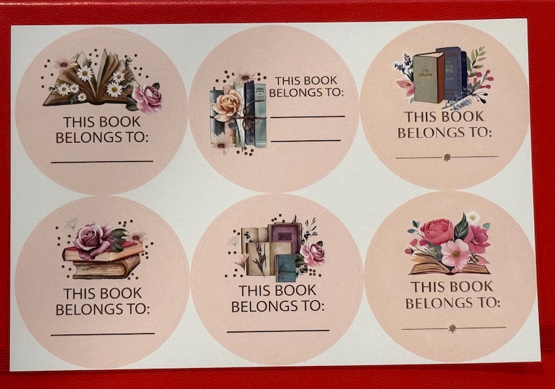 K&ouml;nnte beinhalten: Bogen mit sechs runden Aufklebern mit Blumen- und Buchillustrationen. Jeder Aufkleber tr&auml;gt den Text "THIS BOOK BELONGS TO:" und eine Zeile zum Beschriften. Die Illustrationen zeigen offene B&uuml;cher, B&uuml;cherstapel und Blumengestecke in Rosa und Lila.