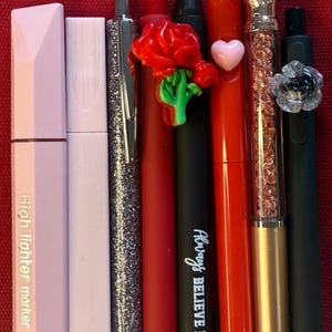 K&ouml;nnte beinhalten: Eine Sammlung von Stiften und Textmarkern in verschiedenen Farben, darunter Rosa, Silber, Rot und Schwarz. Einige Stifte weisen dekorative Elemente wie eine Rose, ein Herz und einen rautenf&ouml;rmigen Aufsatz auf. Ein Stift tr&auml;gt den Text "Always Believe in Yourself".