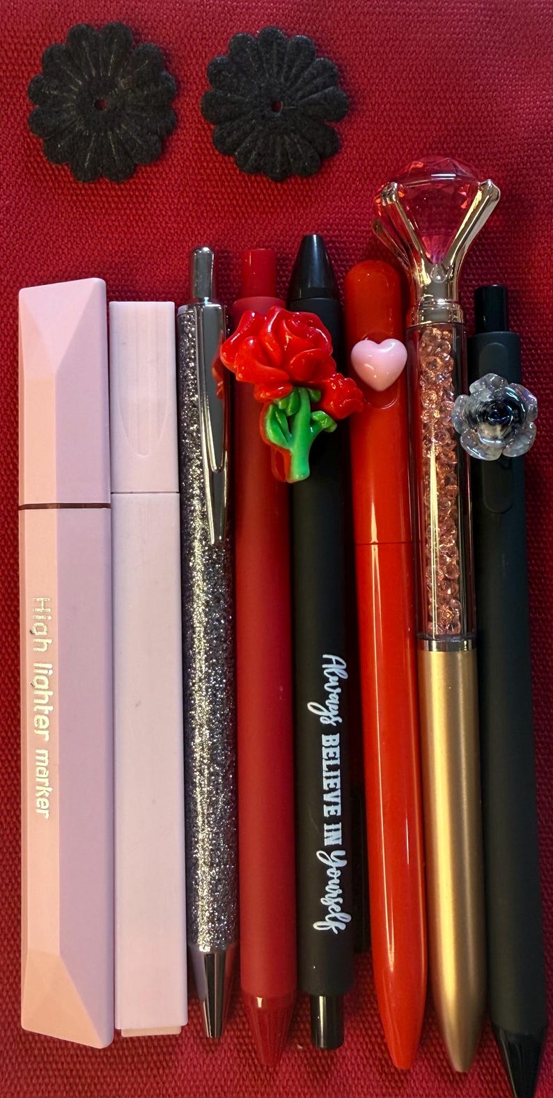 K&ouml;nnte beinhalten: Eine Sammlung von Stiften und Textmarkern in verschiedenen Farben, darunter Rosa, Silber, Rot und Schwarz. Einige Stifte weisen dekorative Elemente wie eine Rose, ein Herz und einen rautenf&ouml;rmigen Aufsatz auf. Ein Stift tr&auml;gt den Text "Always Believe in Yourself".