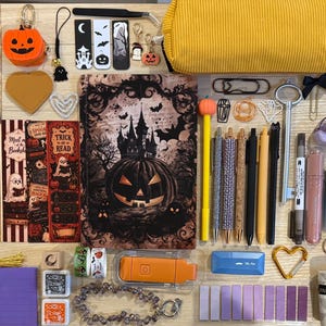 May include: A collection of stationery and office supplies featuring Halloween-themed items, including a notebook with a pumpkin illustration, a yellow pencil case, various pens, bookmarks, stickers, and small decorative charms on a light surface.