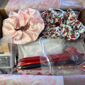 K&ouml;nnte beinhalten: Eine Geschenkbox gef&uuml;llt mit verschiedenen Artikeln, darunter ein gro&szlig;es pinkfarbenes Satin-Haargummi, ein Blumen-Haargummi, Stifte und ein kleiner Stempel. Die Box ist mit Seidenpapier ausgekleidet und enth&auml;lt eine Vielzahl von Accessoires.