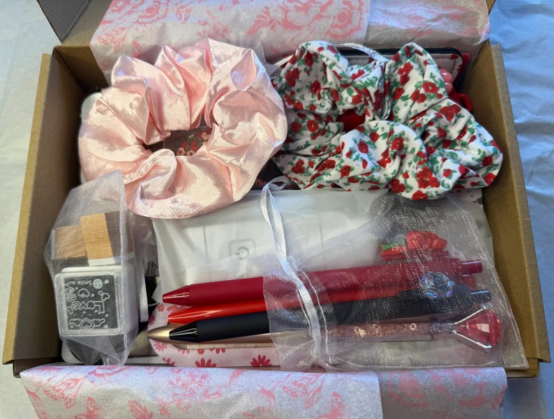 K&ouml;nnte beinhalten: Eine Geschenkbox gef&uuml;llt mit verschiedenen Artikeln, darunter ein gro&szlig;es pinkfarbenes Satin-Haargummi, ein Blumen-Haargummi, Stifte und ein kleiner Stempel. Die Box ist mit Seidenpapier ausgekleidet und enth&auml;lt eine Vielzahl von Accessoires.