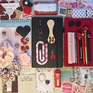 K&ouml;nnte beinhalten: Eine Sammlung von Schreibwaren, darunter ein schwarzes Notizbuch, Stifte, Aufkleber und ein Tagesplaner. Die Artikel sind in Rot-, Rosa- und Wei&szlig;t&ouml;nen gehalten, mit Blumen- und Herzmotiven. Es gibt auch einen roten Tacker und ein rosa Scrunchie.