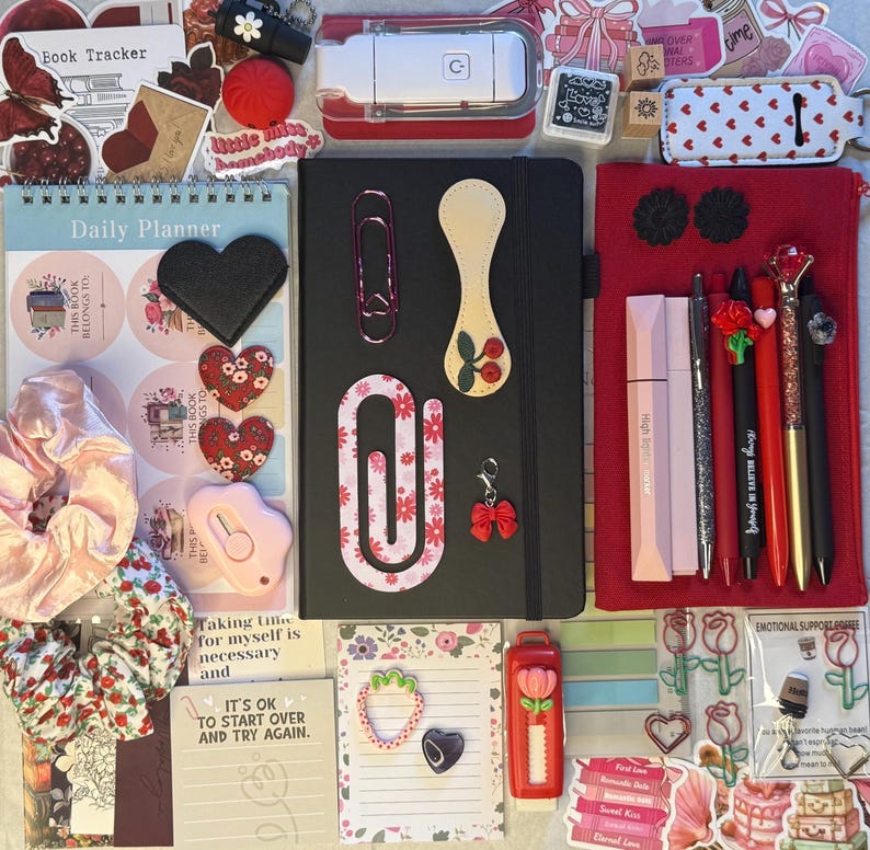 K&ouml;nnte beinhalten: Eine Sammlung von Schreibwaren, darunter ein schwarzes Notizbuch, Stifte, Aufkleber und ein Tagesplaner. Die Artikel sind in Rot-, Rosa- und Wei&szlig;t&ouml;nen gehalten, mit Blumen- und Herzmotiven. Es gibt auch einen roten Tacker und ein rosa Scrunchie.