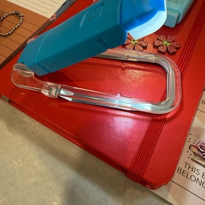 K&ouml;nnte beinhalten: Ein rotes Notizbuch mit Gummiband, einem durchsichtigen Kunststoff-Stifthalter und einem blauen LED-Licht. Das Notizbuch ist von dekorativen Gegenst&auml;nden umgeben, darunter blumenf&ouml;rmige Verzierungen und ein kleines Armband.