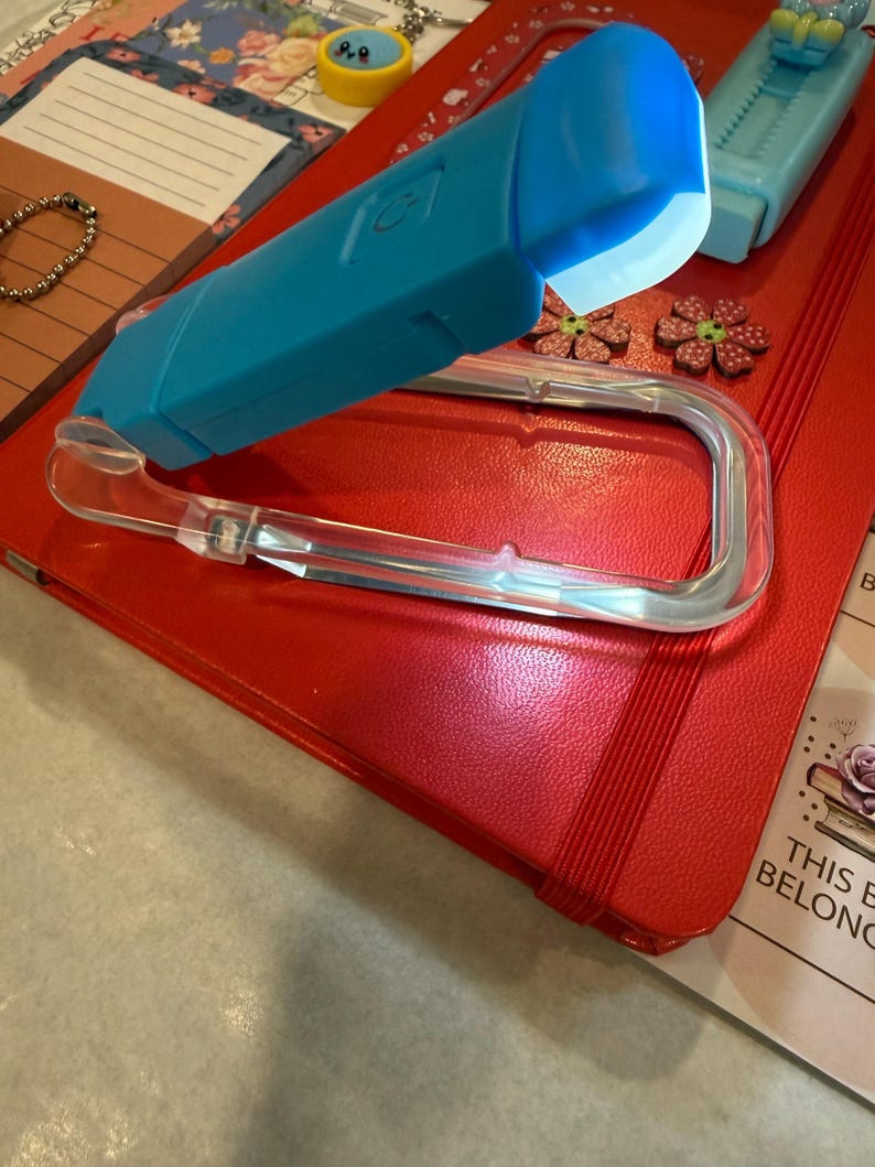 K&ouml;nnte beinhalten: Ein rotes Notizbuch mit Gummiband, einem durchsichtigen Kunststoff-Stifthalter und einem blauen LED-Licht. Das Notizbuch ist von dekorativen Gegenst&auml;nden umgeben, darunter blumenf&ouml;rmige Verzierungen und ein kleines Armband.
