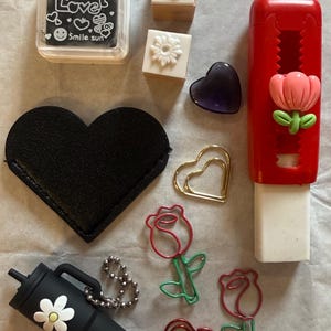 K&ouml;nnte beinhalten: Eine Auswahl an Schreibwaren und Accessoires. Dazu geh&ouml;ren eine schwarze herzf&ouml;rmige Tasche, ein schwarzer Becher mit einer G&auml;nseblume und rosenf&ouml;rmige B&uuml;roklammern. Ebenfalls enthalten sind Stempel, ein lila Herz und ein roter Radiergummi.