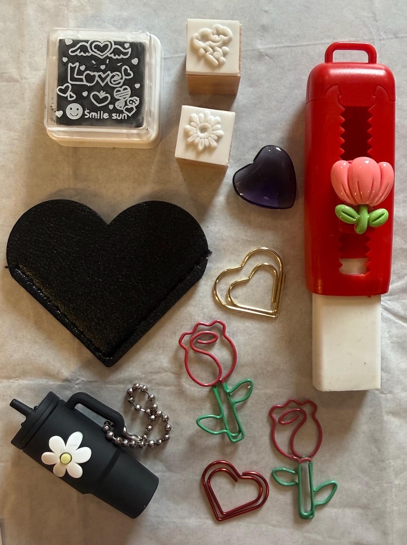 K&ouml;nnte beinhalten: Eine Auswahl an Schreibwaren und Accessoires. Dazu geh&ouml;ren eine schwarze herzf&ouml;rmige Tasche, ein schwarzer Becher mit einer G&auml;nseblume und rosenf&ouml;rmige B&uuml;roklammern. Ebenfalls enthalten sind Stempel, ein lila Herz und ein roter Radiergummi.