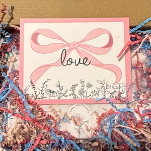 K&ouml;nnte beinhalten: Eine rosa Karte mit einer rosa Schleife und dem Wort "love" in Schreibschrift. Die Karte ist mit einem Blumendesign am unteren Rand verziert und von farbigem Zerkleinerungspapier in einem Karton umgeben.