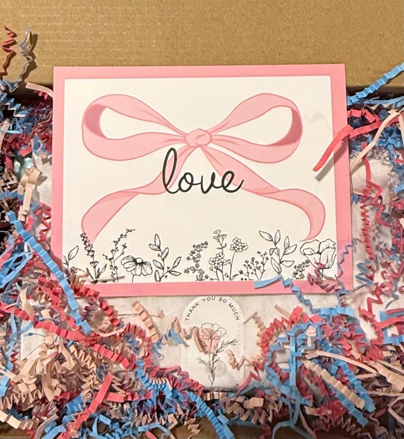 K&ouml;nnte beinhalten: Eine rosa Karte mit einer rosa Schleife und dem Wort "love" in Schreibschrift. Die Karte ist mit einem Blumendesign am unteren Rand verziert und von farbigem Zerkleinerungspapier in einem Karton umgeben.