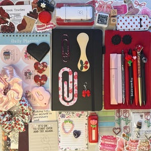 K&ouml;nnte beinhalten: Eine Sammlung von Schreibwaren, darunter ein schwarzes Notizbuch, Stifte, Aufkleber und ein Tagesplaner. Die Artikel sind in Rot-, Rosa- und Schwarzt&ouml;nen gehalten und mit Herz- und Blumendesigns versehen. Es gibt auch B&uuml;roklammern, einen Schleifenanh&auml;nger und eine rosa Haargummi.